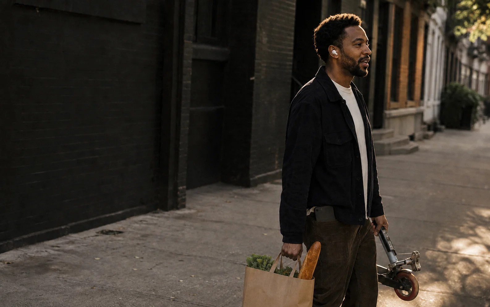 A man walks with a grocery bag and folded scooter in both hands, speaking through one earbud while his phone remains in his back pocket.